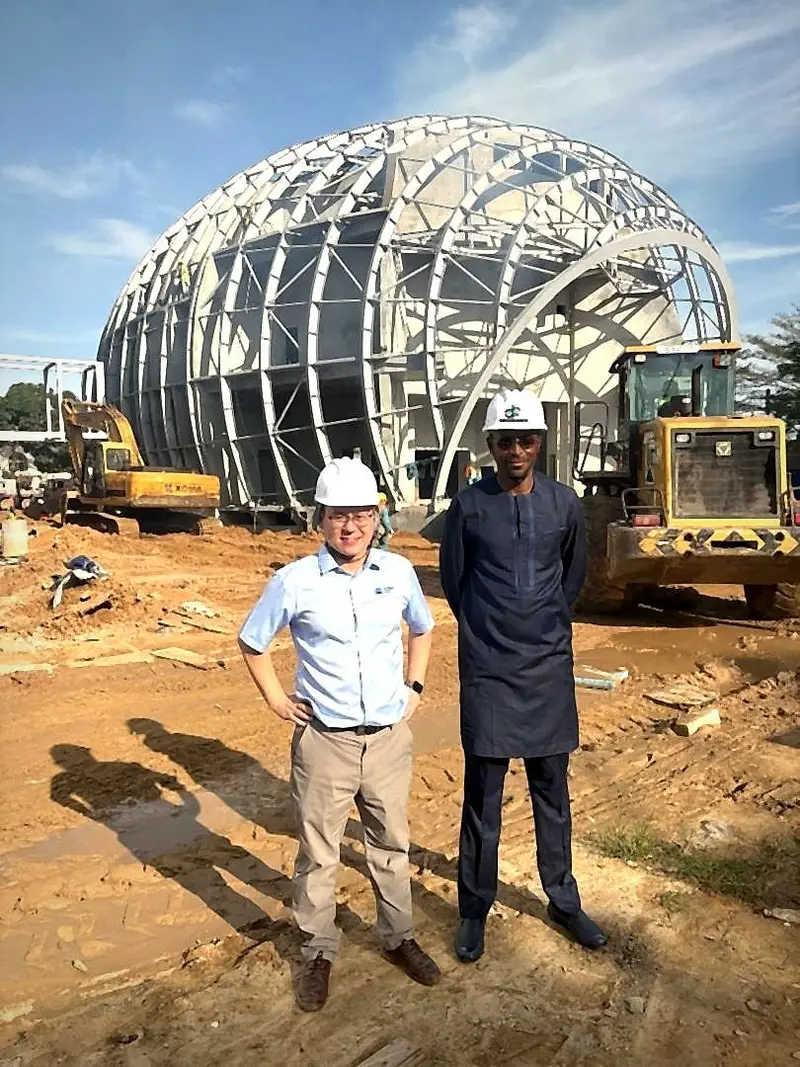Deux responsables en casque de chantier blanc devant une grande structure métallique en forme de dôme, sur un chantier avec des engins de travaux